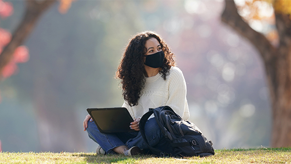 Female student sitting on the ground, laptop on lap, backpack next to her, with a mask on.