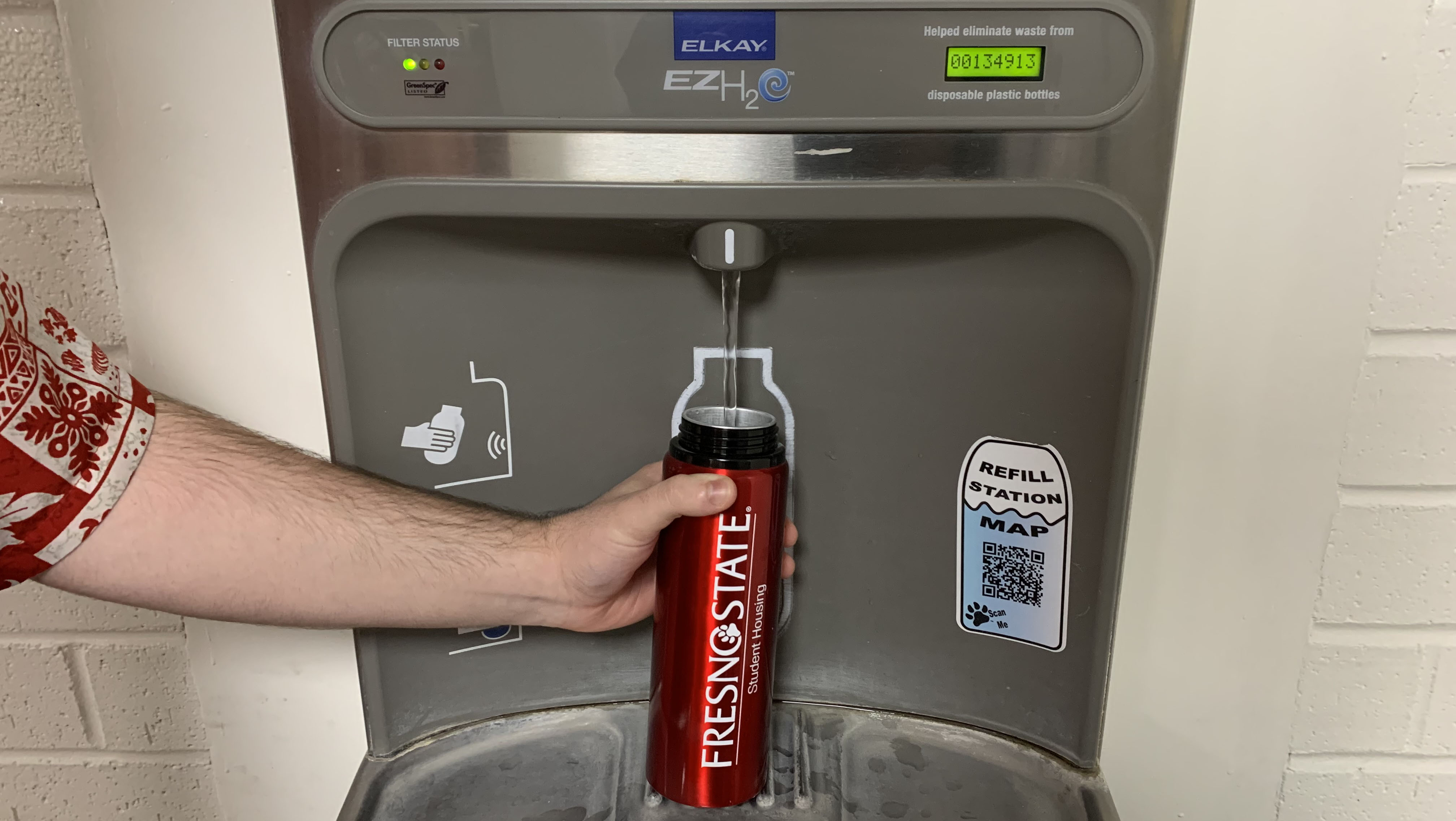 Mans hand holding a red water bottle while filling it at one of the water bottle filling stations on campus.