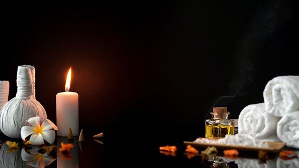 A lit white candle, rolled up hand towels and flower petals against a black background