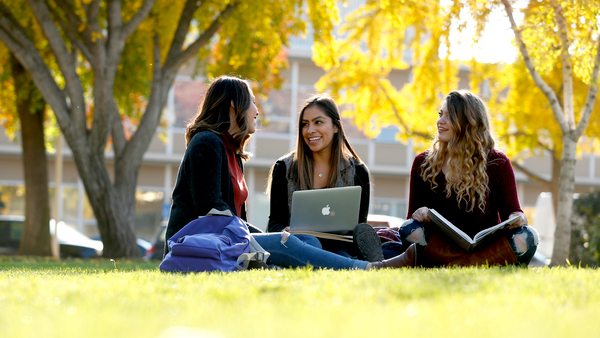 Three female students sitting on the grass talking with yellow fall trees in the background