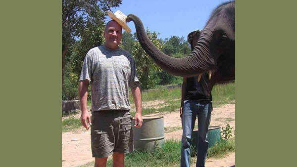 Dr. Steve Blumenshine standing next to an elephant who is using its trunk to lift a straw hat off or onto Blumenshine