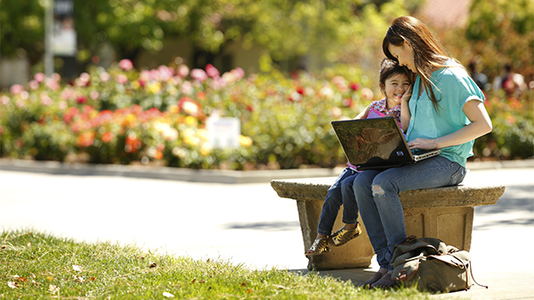 Woman sitting on a bench with a laptop and her young daughter leaning on her