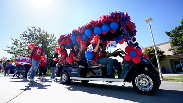 Students sit in a decorated golf cart with blue and red balloons waving red pom poms.