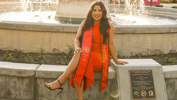 Audrey Munoz posing in front of the Fresno State fountain in a red dress with a graduation sash on.