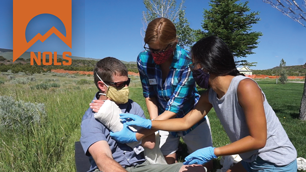 A woman gives first aid to a main with a bandaged hand in a meadow. NOLS graphic in the upper left corner.