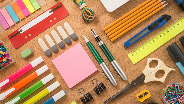 A desk with office supplies like highlighters, sticky notes, a ruler, scissors, pencils and paper clips.