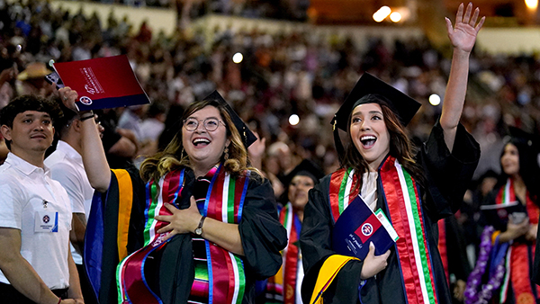 Two Hispanic women graduates smiling and raising arms as they graduate.