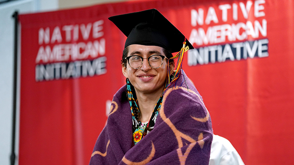 Native American student wrapped in a blanket in front of a banner that reads Native American Initiative