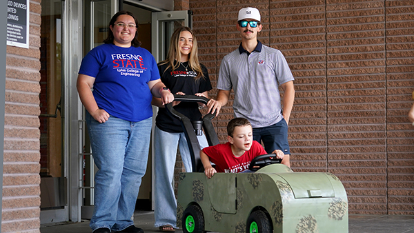 Beau Bewarder sits in a camouflage green toy car in front of three Fresno State students.