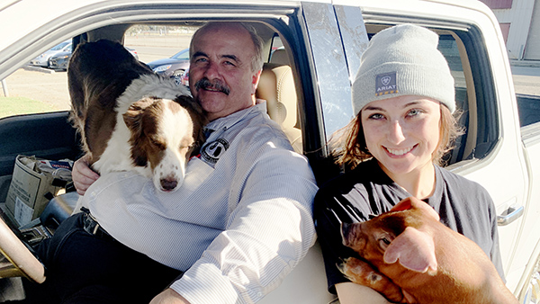 Dr. Scott Williamson sits in the driver seat of a white truck with a dog leaning over his shoulder. Daughter, Hannah, leans against the truck holding a baby pig.