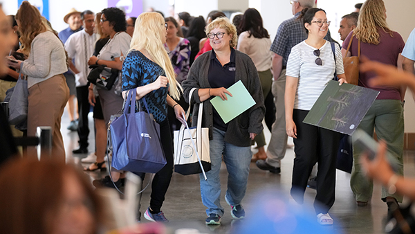 A crowd of people surrounding two women talking to each other, holdings bags.