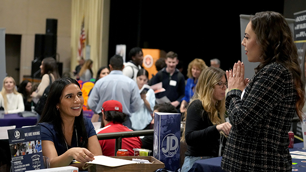One woman seated at a table and another stands in front of her talking.