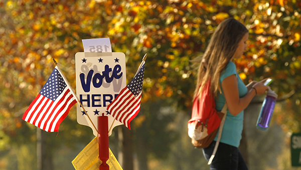 A girl on her phone walking past a Vote Here sign.