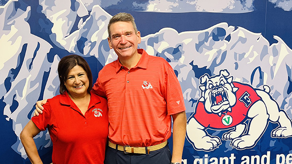 Belinda Mu&ntilde;oz, admin support coordinator for the Center for Irrigation Technology, (left), and Garrett Klassy, Director of Athletics, (left), standing in front of a wall with blue mountains and sky and the Fresno State bulldog on the right side of the photo.